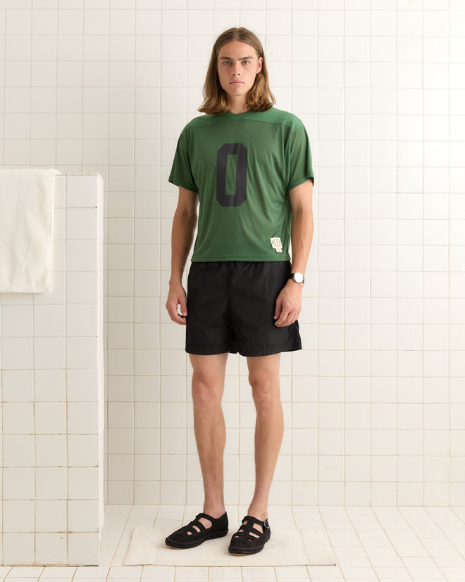 A man wearing the Scrimmage Jersey - Green with a large "0" and black shorts stands in a white tiled room.