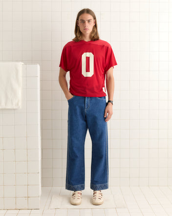 A person stands in a tiled room wearing the Scrimmage Jersey - Red, wide jeans, and white sneakers.