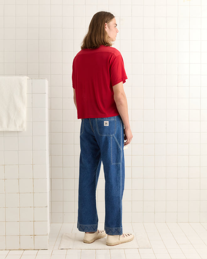 A person wearing the Scrimmage Jersey - Red and blue jeans stands facing a white tiled wall in a minimalist room.