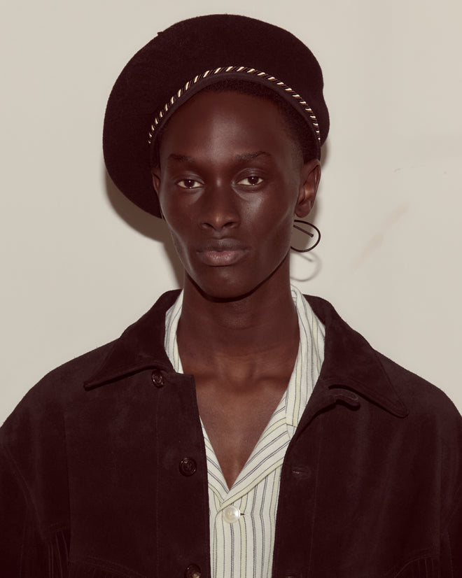 A person wears the Script Beret in black with brown/black piping and a jacket over a striped shirt, looking at the camera.