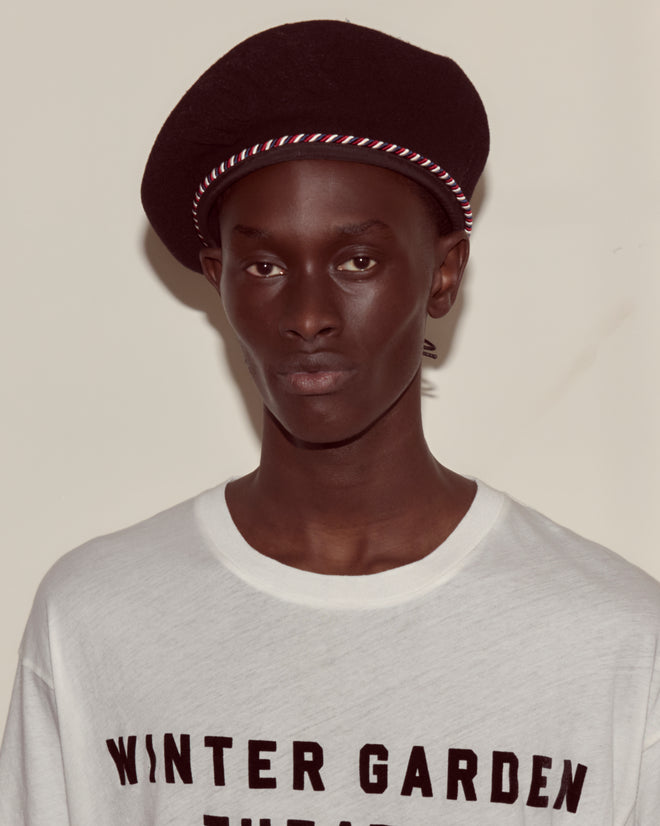 A person wearing the Script Beret - Black with Red/Blue Piping and a white "WINTER GARDEN PUB" shirt stands against a plain background.