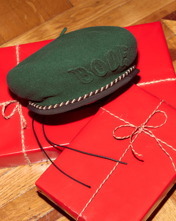 A Script Beret in green with brown/black piping rests on two red, gift-wrapped boxes tied with twine bows.