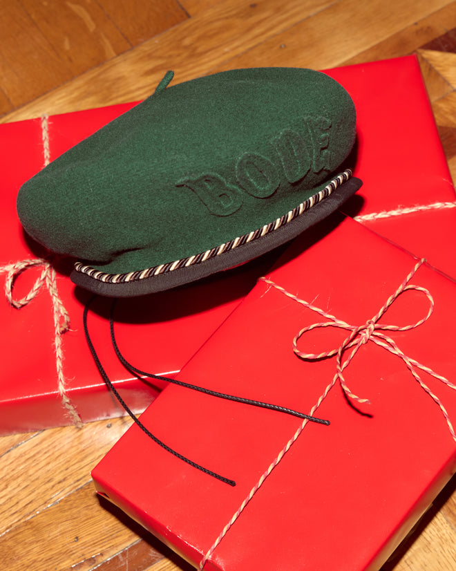 A Script Beret in green with brown/black piping rests on two red, gift-wrapped boxes tied with twine bows.
