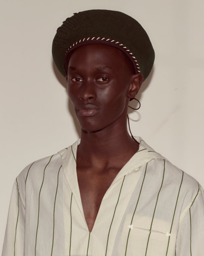A person wearing a striped shirt and the Script Beret - Green with Brown/Black Piping, made in France from 100% wool, stands against a plain background, looking at the camera.