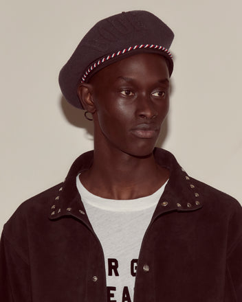 A person wearing the Script Beret in charcoal with red/blue piping and a jacket with metal studs over a white printed shirt, looking to the side.