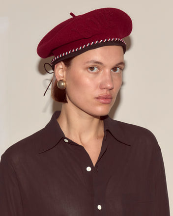 Woman wearing the Script Beret - Bordeaux with Red/Blue Piping, made in France, and large round earrings, looks at the camera in a dark shirt.