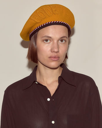 Woman wearing the Script Beret in gold with red and blue piping, styled like a classic French beret and paired with a dark button-up shirt, looking at the camera.