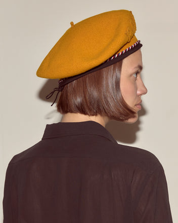 Woman in a Script Beret - Gold with Red/Blue Piping and a dark shirt faces sideways against a plain background.