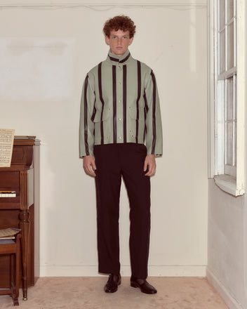 Indoors, a man stands by a piano wearing the Seafoam Stripe Welding Jacket in seafoam/black paired with black pants.