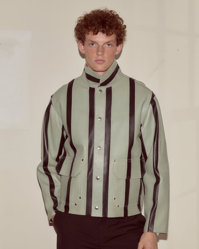 A person with curly hair wears the Seafoam Stripe Welding Jacket in seafoam and black, featuring bold black vertical stripes, while standing against a plain wall.