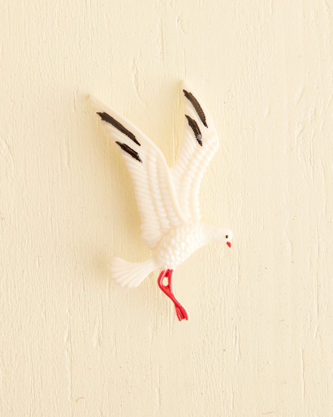 A Seagull Brooch showcasing a vintage design with a white plastic silhouette of a seagull, complete with black-tipped wings and red beak and legs, affixed to a light-colored textured wall.