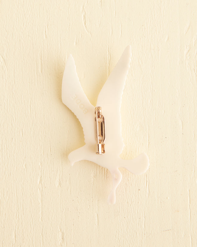 A Seagull Brooch showcasing a vintage design with a white plastic silhouette of a seagull, complete with black-tipped wings and red beak and legs, affixed to a light-colored textured wall.