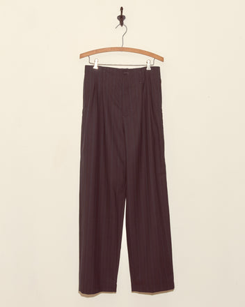 Sea Levels Trousers - 28 in dark pinstriped deadstock wool hang on a wooden hanger against a plain off-white wall.