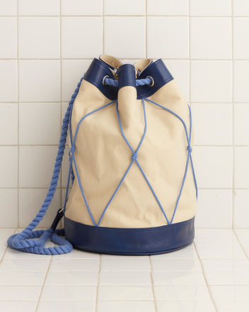 The Sea Rig Rucksack, a heavyweight canvas drawstring bag in beige and blue, with a braided rope strap, elegantly rests on a tiled surface.