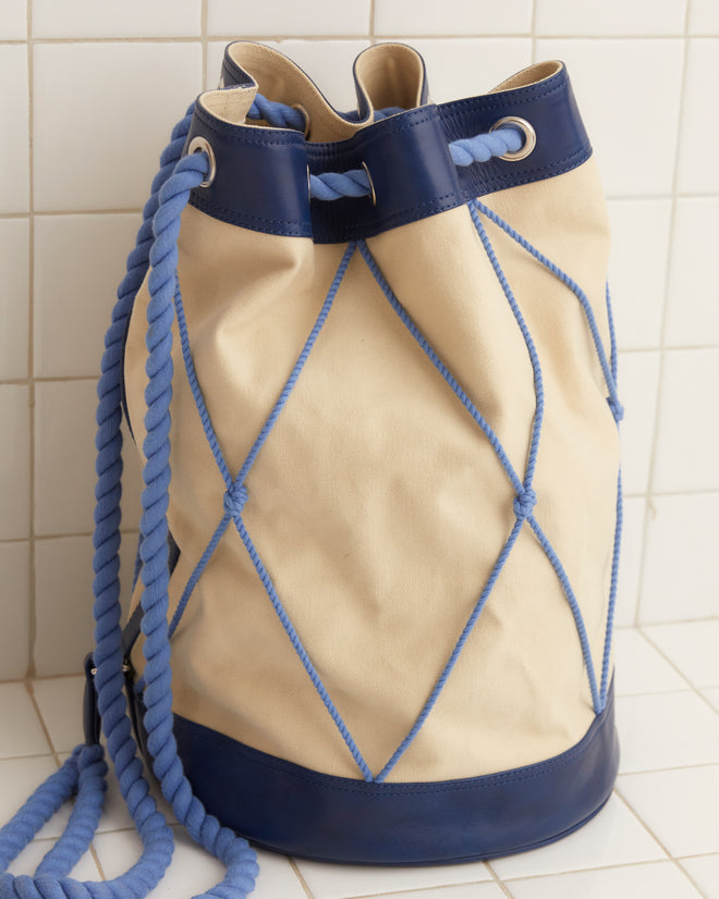 The Sea Rig Rucksack, a heavyweight canvas drawstring bag in beige and blue, with a braided rope strap, elegantly rests on a tiled surface.