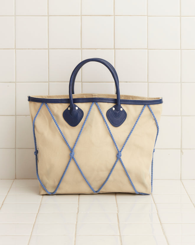 The Sea Rig Tote Bag, made from durable tan canvas and accented with blue leather details and a diamond pattern, is showcased on a white tiled background.