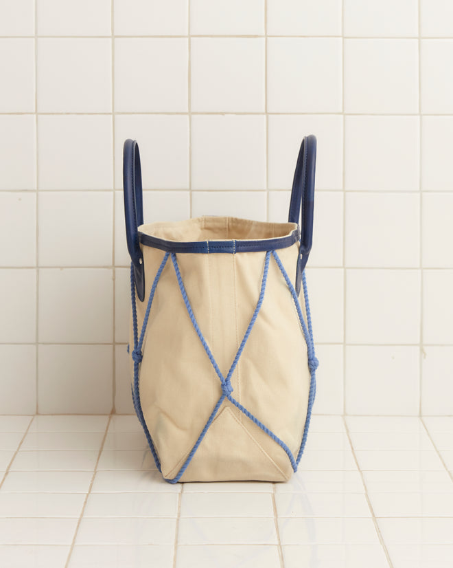 The Sea Rig Tote Bag, made from durable tan canvas and accented with blue leather details and a diamond pattern, is showcased on a white tiled background.