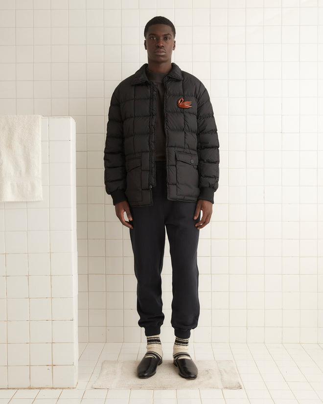 A man stands in a tiled room wearing the Seattle Puffer Jacket in black and dark pants with slip-on shoes. A towel hangs on the left, complementing his classic American outdoor wear vibe.