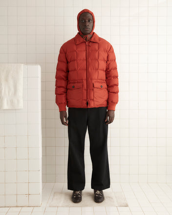 A person wearing the Seattle Puffer Jacket in Brick, featuring a nylon shell and hood, stands against a white tiled wall in a minimal setting.