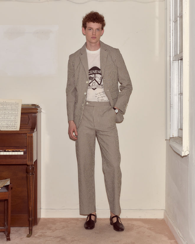 A person wearing the Seersucker Cafe Stripe Suit Jacket with a notched lapel and a graphic T-shirt stands by a piano in a sparsely furnished room with white walls.