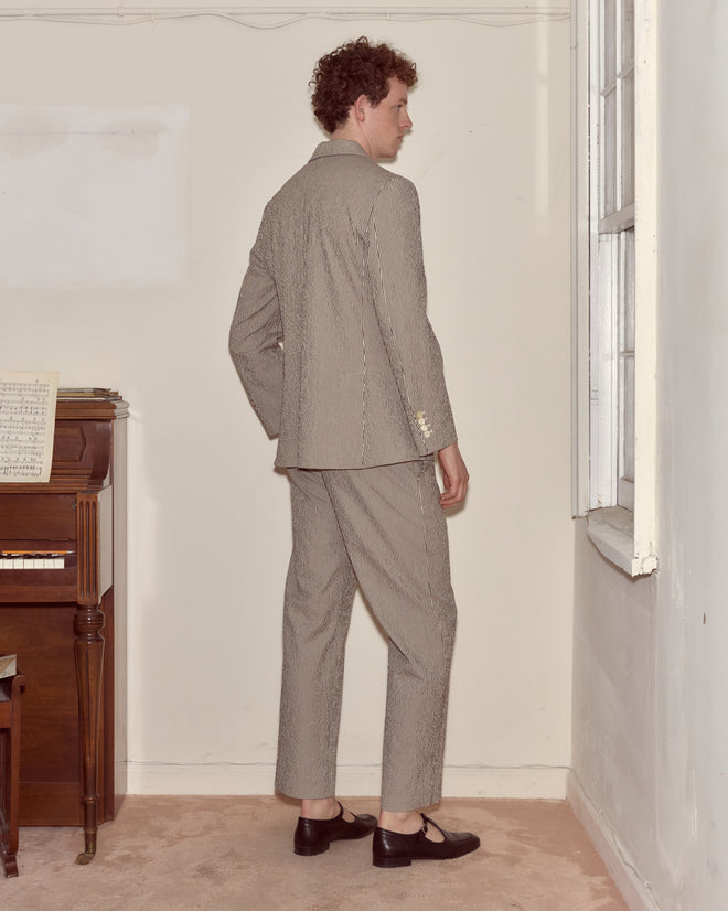 A man in a Seersucker Cafe Stripe Suit Jacket with a notched lapel stands by a window, facing sideways, near a piano with sheet music.