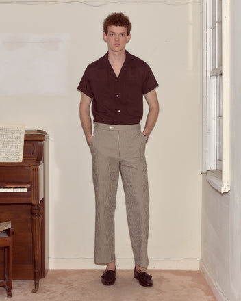 A man in a maroon short-sleeve shirt and Seersucker Cafe Stripe Trousers stands near a piano in a light-colored room.