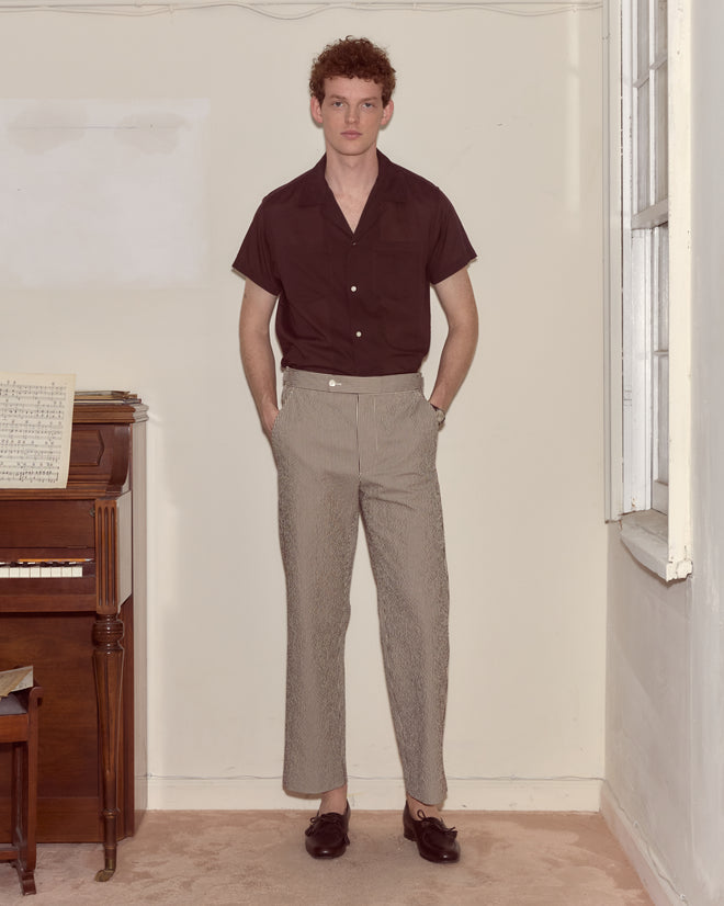 A man in a maroon short-sleeve shirt and Seersucker Cafe Stripe Trousers stands near a piano in a light-colored room.