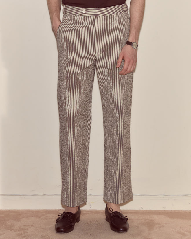 Man wearing Seersucker Cafe Stripe Trousers, black shoes, and a watch, standing with one hand in his pocket against a plain background.