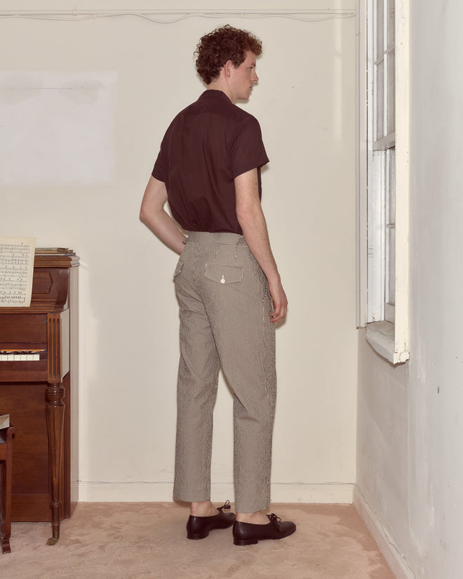 A man in a brown shirt and Seersucker Cafe Stripe Trousers stands near a window, facing away from the camera.