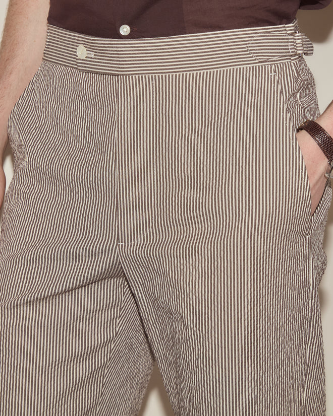 Person wearing Seersucker Cafe Stripe Trousers with brown and white stripes, straight leg fit, hands in pockets, and a partially visible brown shirt.