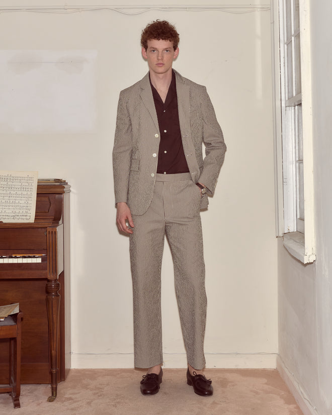 A man in a tan suit with a dark shirt and Seersucker Cafe Stripe Trousers stands indoors by a piano and window, looking at the camera.