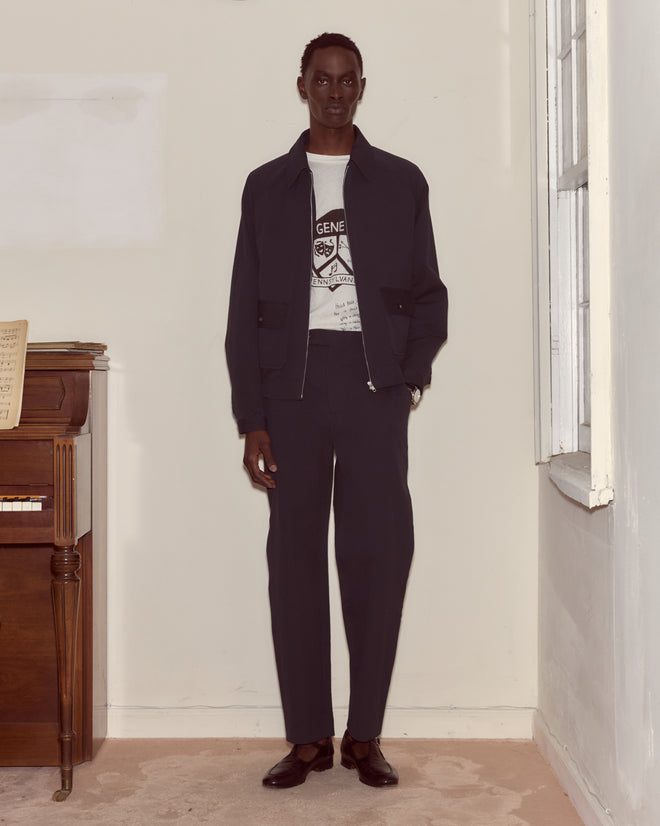 A man in a dark suit and graphic t-shirt stands indoors by a piano and window, layering with the Seersucker Coach's Jacket for laid-back style.