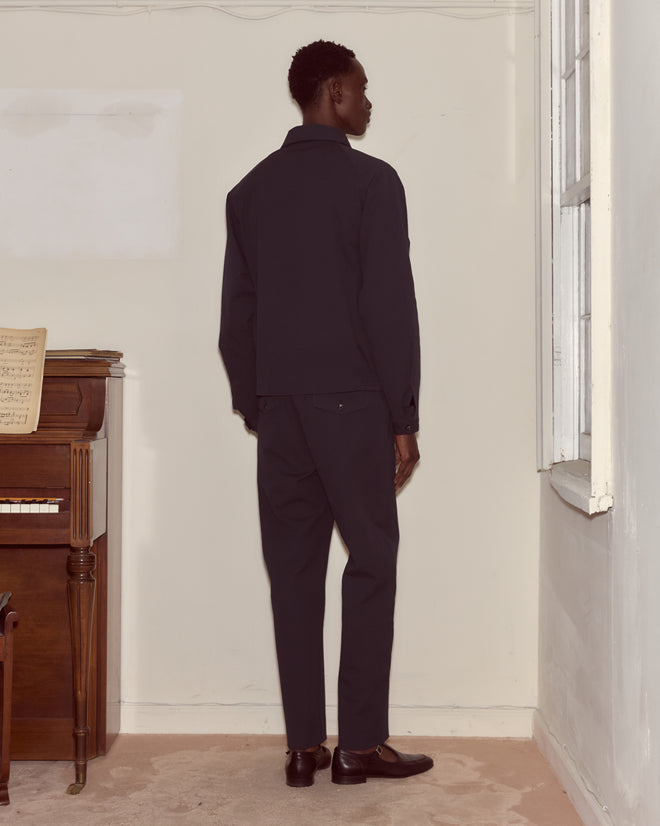 A person wearing a Seersucker Coach's Jacket stands facing the corner of a room near a piano and a window.