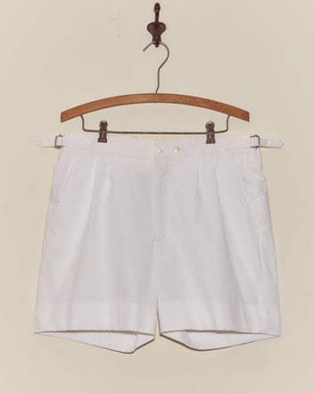 Seersucker Double-Button Shorts hang on a wooden hanger against an off-white wall, showcasing their crisp pleated design with side adjusters for a vintage style look.
