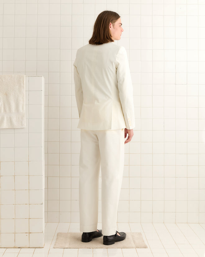 A person with long hair, dressed in a Seersucker Double-Breasted Suit Jacket, stands facing a tiled wall in a minimalist room.
