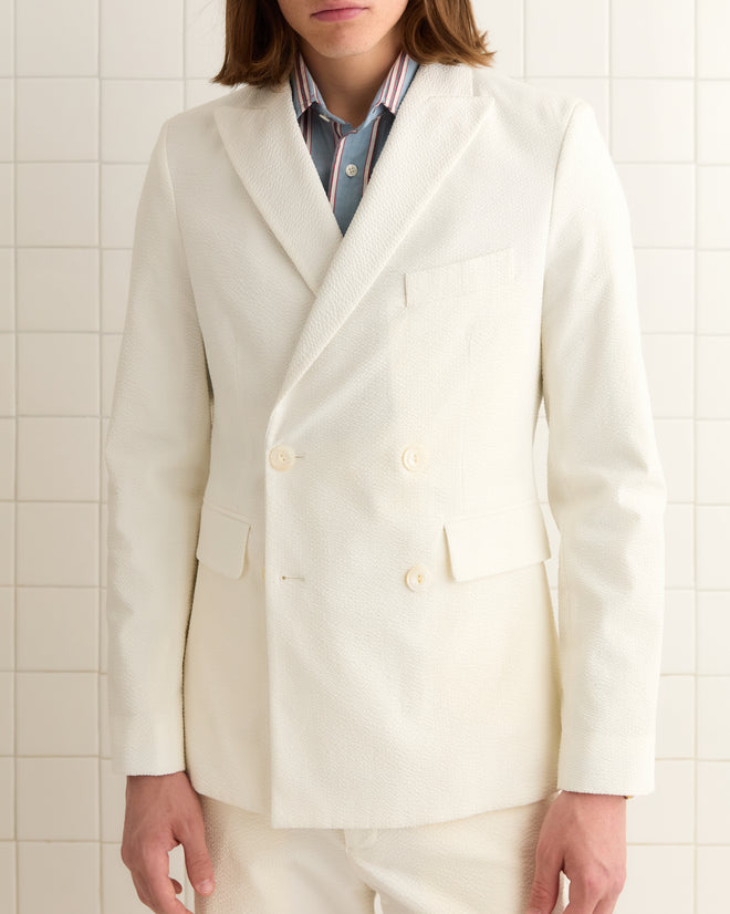 Wearing the Seersucker Double-Breasted Suit Jacket, a person stands confidently against a tiled wall. The sleek white suit jacket is paired with a striped shirt, featuring distinctive double-breasted buttons.