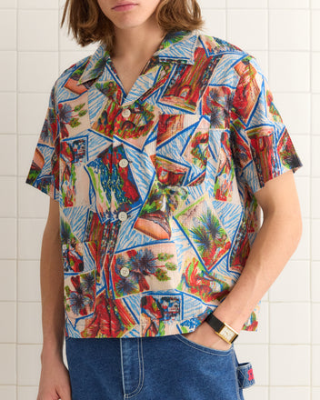 Clad in a Seersucker Postcard Short Sleeve Shirt and blue jeans, a person stands against a tiled wall, effortlessly blending style and comfort.