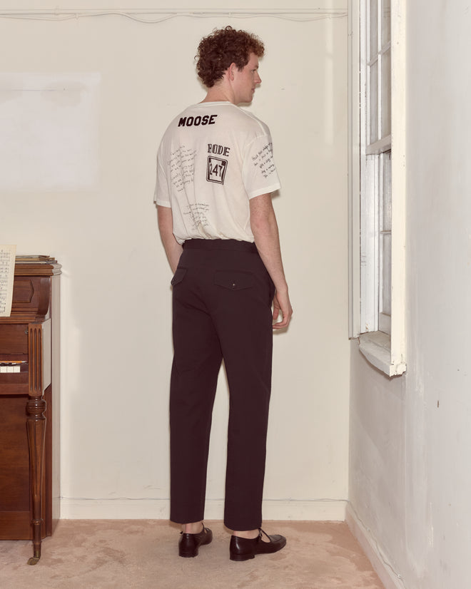 A man stands indoors facing away, wearing a graphic T-shirt, Seersucker Side Buckle Trousers - Dark Navy, and black shoes.