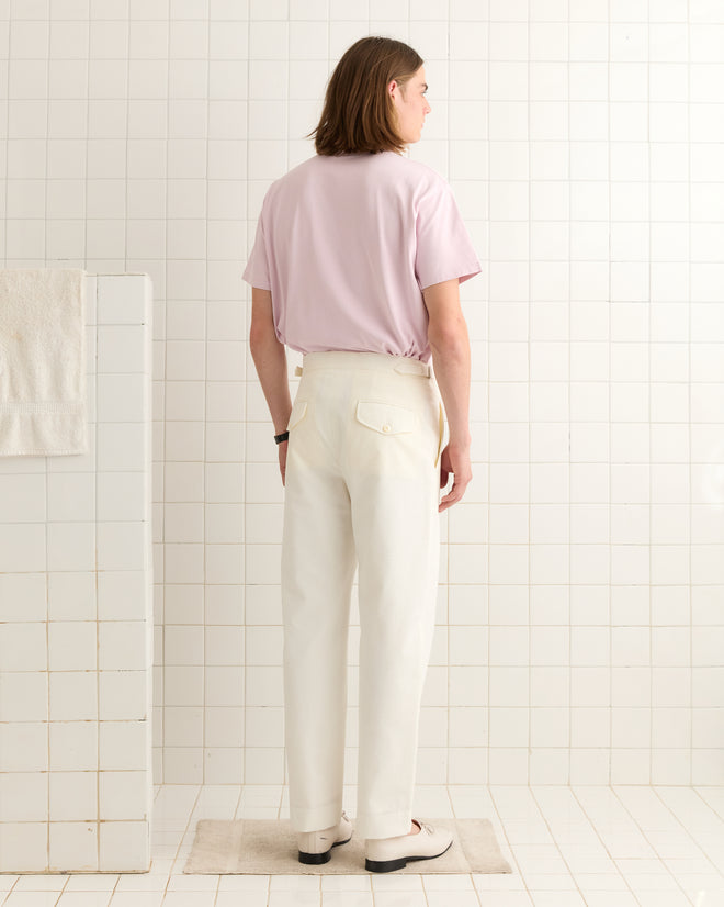 A person with long hair, dressed in a pink shirt and Seersucker Suiting Trousers styled in vintage men's fashion with mid-rise straight leg pants, stands in a tiled room facing away.
