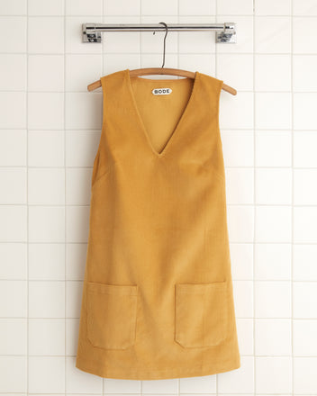 The Custom Senior Cord Dress in mustard hangs elegantly on a hanger against a backdrop of white tiled walls, reminiscent of the classic British corduroy style.