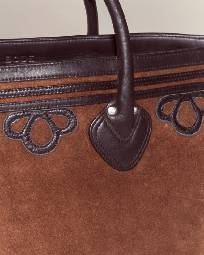 The Trefoil Tote - Brown features a boxy silhouette, brown suede body, black leather handles, and decorative black leather accents for a modern look.