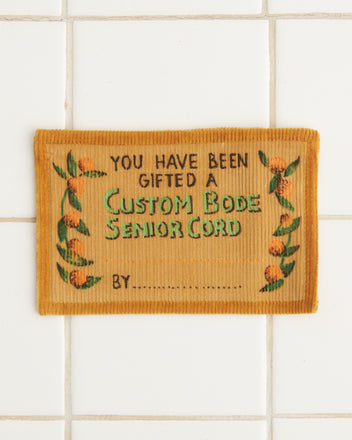 A British corduroy patch is set against a tiled background embellished with floral designs and elegant text detailing the Custom Senior Cord Gift Certificate.