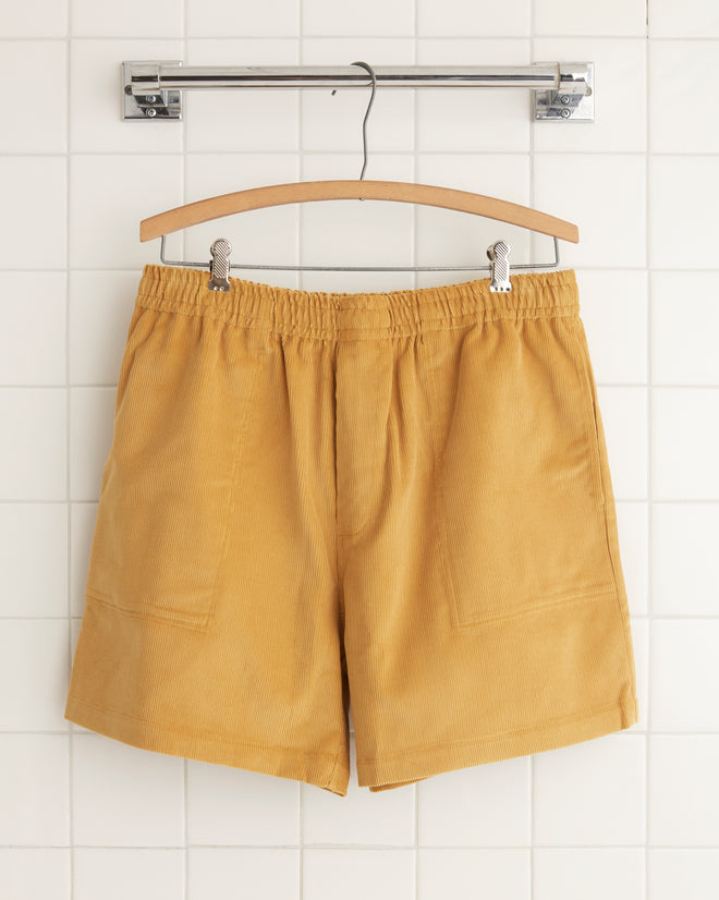 The Custom Senior Cord Shorts in yellow hang on a wooden hanger against a tiled wall.