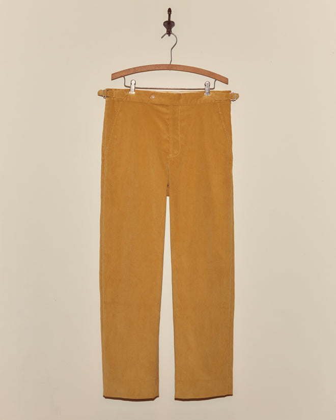 A pair of Custom Senior Cord Side-Buckle Trousers in mustard yellow British corduroy hang on a wooden hanger against a beige wall.