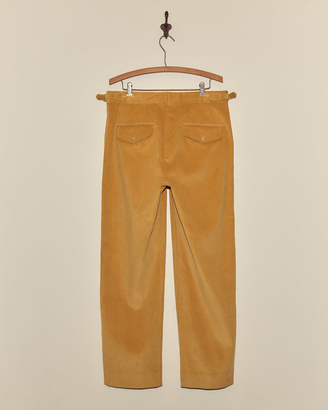 Custom Senior Cord Side-Buckle Trousers in yellow corduroy hang from a wooden hanger against a plain beige wall.