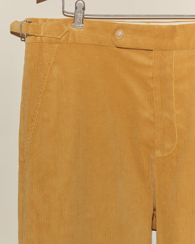 Close-up of Custom Senior Cord Side-Buckle Trousers in mustard yellow British corduroy, displayed on a metal hanger against a beige background.