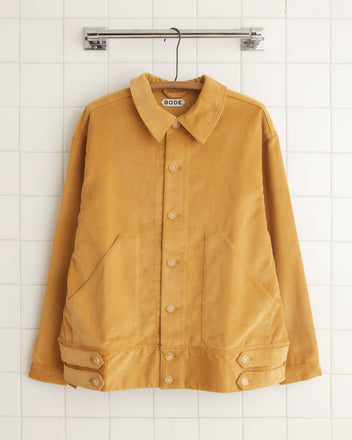 A Custom Senior Cord Side Tab Jacket in tan, featuring several buttons, a collar, and front pockets made of British corduroy fabric, hangs on a wooden hook against a beige wall. The label 