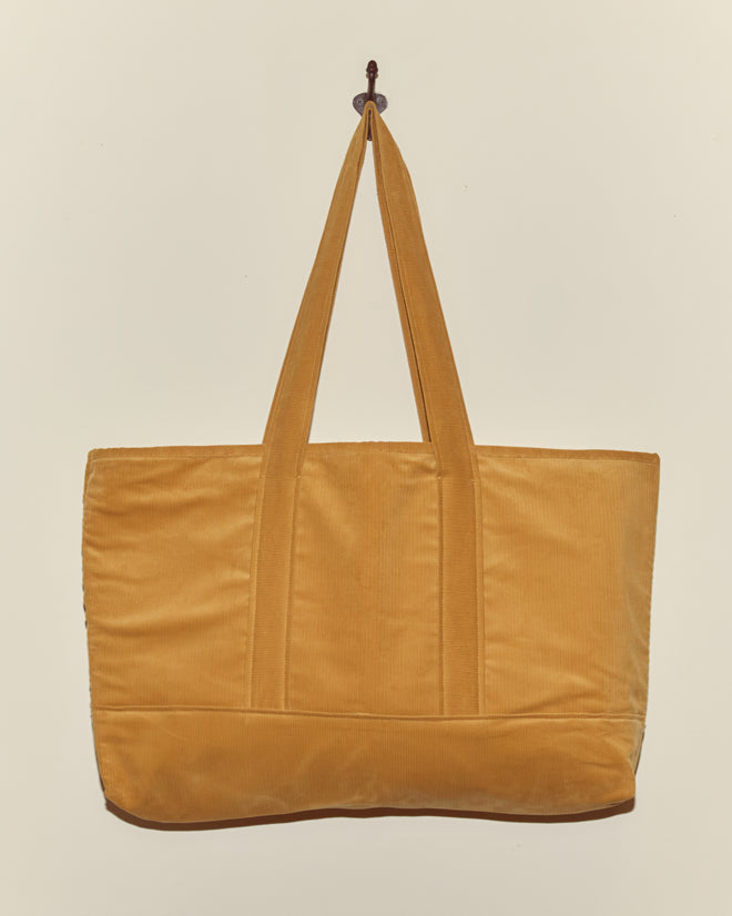 The Custom Large Senior Cord Tote in mustard yellow, with two handles, hangs on a hook against a light background—ideal for carrying your corduroy notebook or daily essentials.