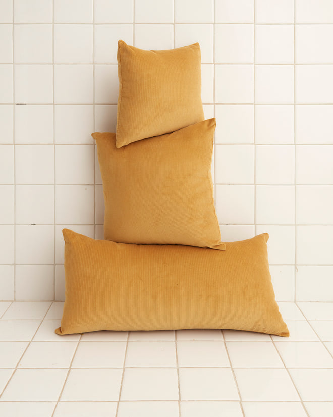 Three mustard-colored Custom Senior Cord pillows are stacked against a white tiled wall and floor, showcasing the elegance of bespoke textiles.