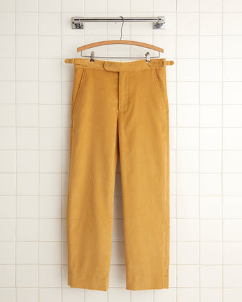 A pair of Custom Senior Cord Side-Buckle Trousers in light brown, featuring pockets, is hanging on a wooden hanger against a white wall with hooks.